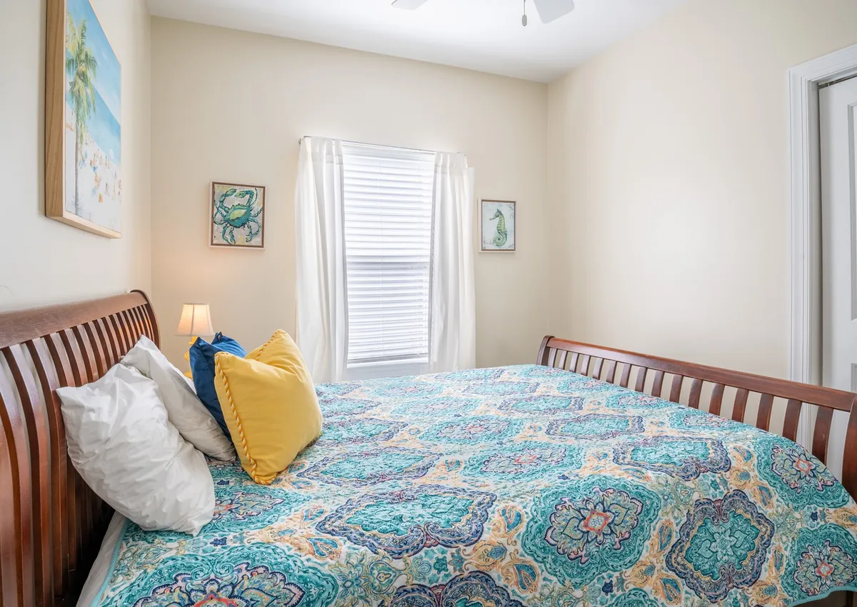A welcoming bedroom with a bed dressed in a colorful quilt, decorative wall art, and a window adorned with sheer curtains. The setting is bright and cheerful, evoking a peaceful retreat perfect for guests or couples.