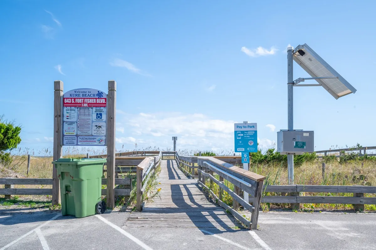 A coastal entrance scene featuring a wooden walkway leading toward the beach, with a relaxed 'Welcome' sign, a 'Pay to Park' sign, green trash bin, and a solar panel. The setting feels welcoming and is instantly recognizable as a gateway to a family beach adventure.