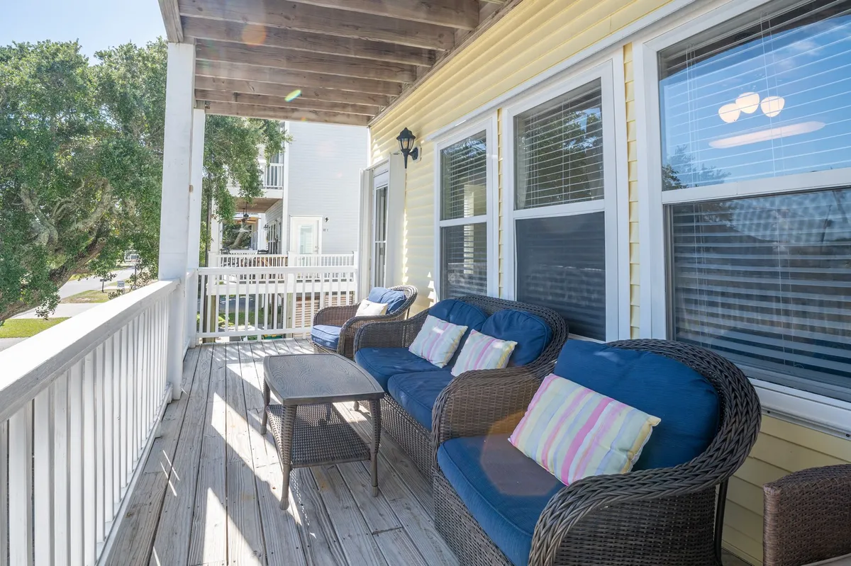 A covered wooden porch featuring wicker seating with colorful cushions and a wooden deck, ideal for relaxing and socializing. The area is designed for comfort and leisure, offering a welcoming transition between indoors and outdoors while overlooking a peaceful street setting.