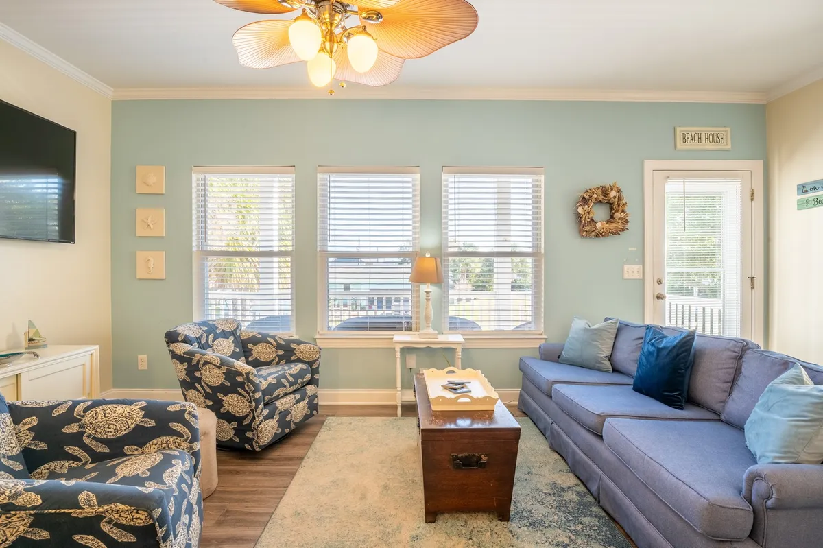 The image showcases a cozy living room featuring a blue sofa, decorative chairs, a coffee table, and large windows that fill the space with natural light. The scene suggests comfort and a welcoming atmosphere, with views that could hint at a connection to the front porch or the peaceful outdoors.