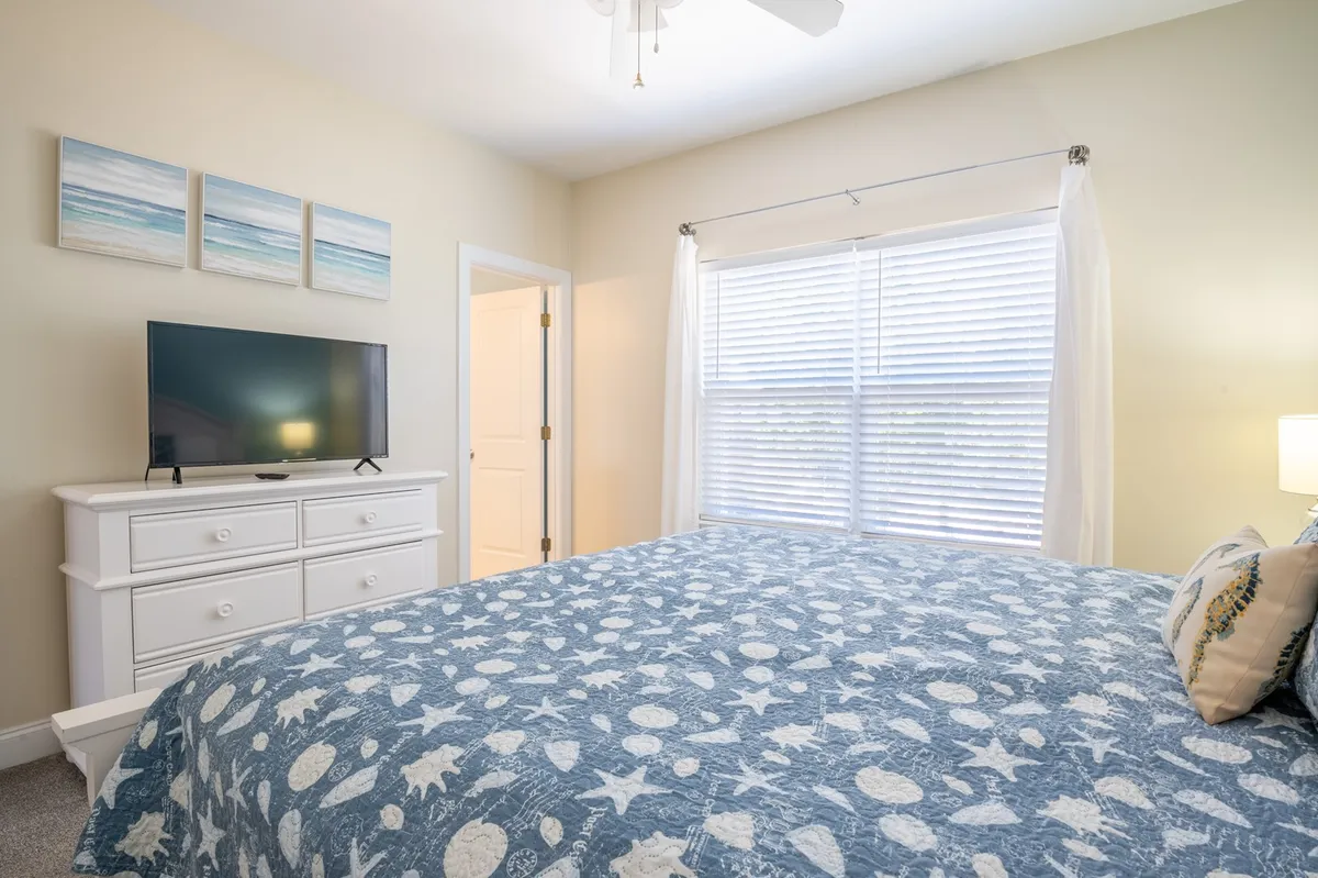 This image features a serene bedroom with a bed covered in blue patterned linens, a television placed on a dresser, a large window with sheer curtains letting in natural light, and beach-themed wall art. The space feels tranquil and inviting, highlighting both comfort and modern amenities.