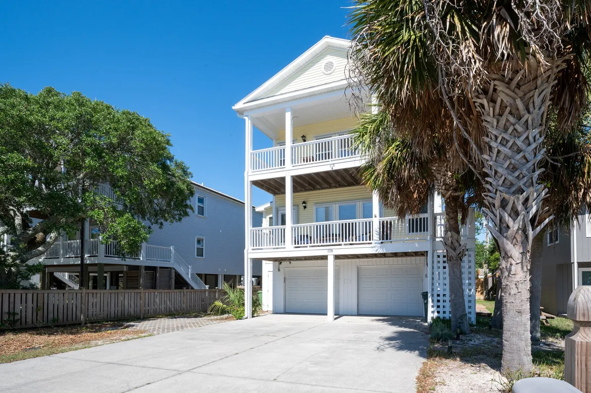 The image showcases the condo's elevated structure framed by a towering palm tree and a spacious, cushioned front porch. The porch provides a welcoming space for gatherings, framed by the gentle glow of sunset and the lush surroundings of Kure Beach. The overall mood is tranquil and inviting, perfect for relaxing with loved ones or enjoying quiet mornings by the ocean breeze.
