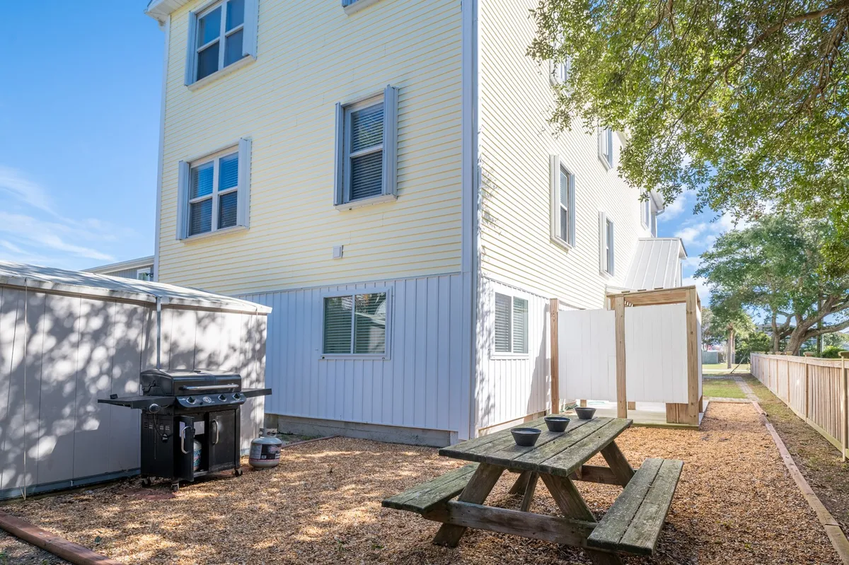 A casual, inviting backyard scene showing a wooden picnic table and a grill set beside a wooden fence, with a shed in the background. The area feels enclosed and safe, ideal for pets and owners to enjoy meals or relax outdoors. Trees provide shade and a sense of privacy.