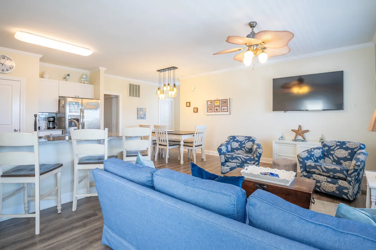 A bright and inviting open-concept living and dining area featuring a plush blue sofa, a farmhouse-style dining table, a ceiling fan with a light fixture, and a wall-mounted smart TV. The space is styled for relaxation and group gatherings, with ample seating and natural light filling the room.