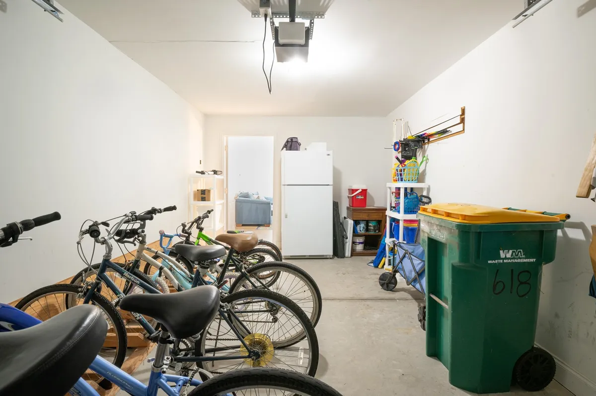 A practical interior shot of a garage area, with multiple bikes stored alongside a refrigerator, green trash bin, and storage shelves. The space is organized and functional, suitable for storing beach gear, bikes, and even a rental golf cart.