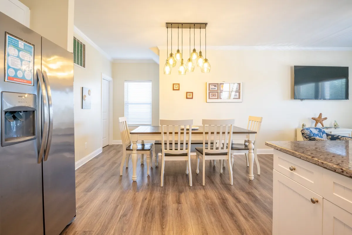 A view of the dining area featuring a farmhouse-style table set beneath a stylish hanging light fixture. The space is warm and welcoming, with the refrigerator and a TV visible in the background, connecting the dining and kitchen zones for a seamless, sociable layout.
