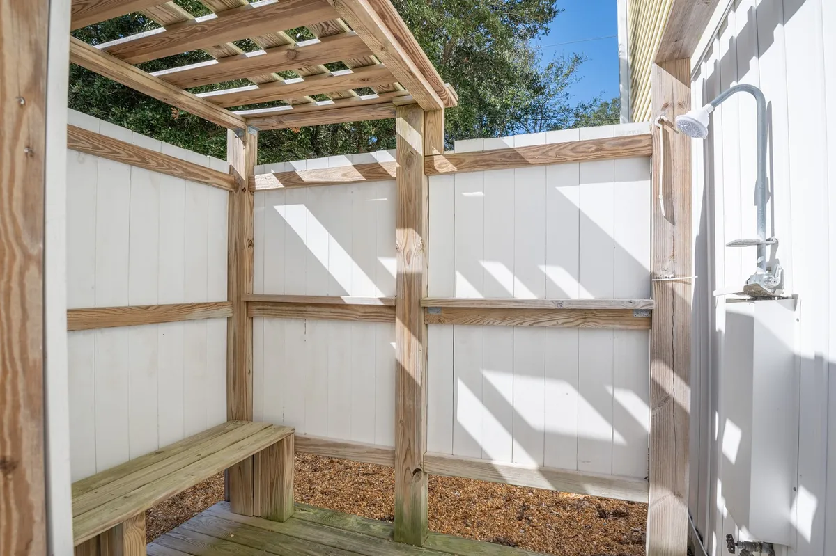 An exterior view showcasing a wooden bench beneath a pergola structure and a shower fixture, indicating a dedicated outdoor shower area. The space is designed for rinsing off after a day at the beach and relaxing in a natural, refreshing environment.