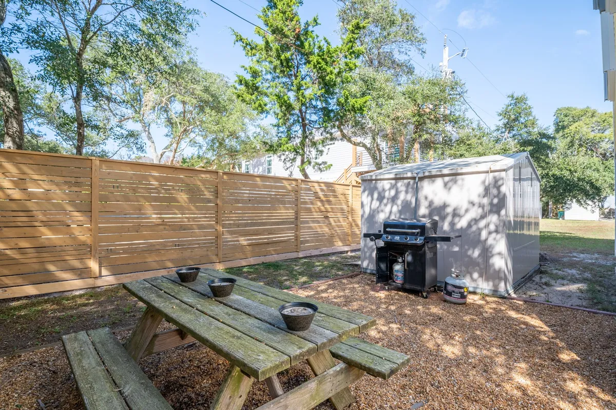 A relaxed, inviting backyard scene featuring a picnic table, a gas grill, a storage shed, and a fenced perimeter. The area is spacious, well-kept, and designed for outdoor dining, gatherings, and barbecues, with room for pets to roam.